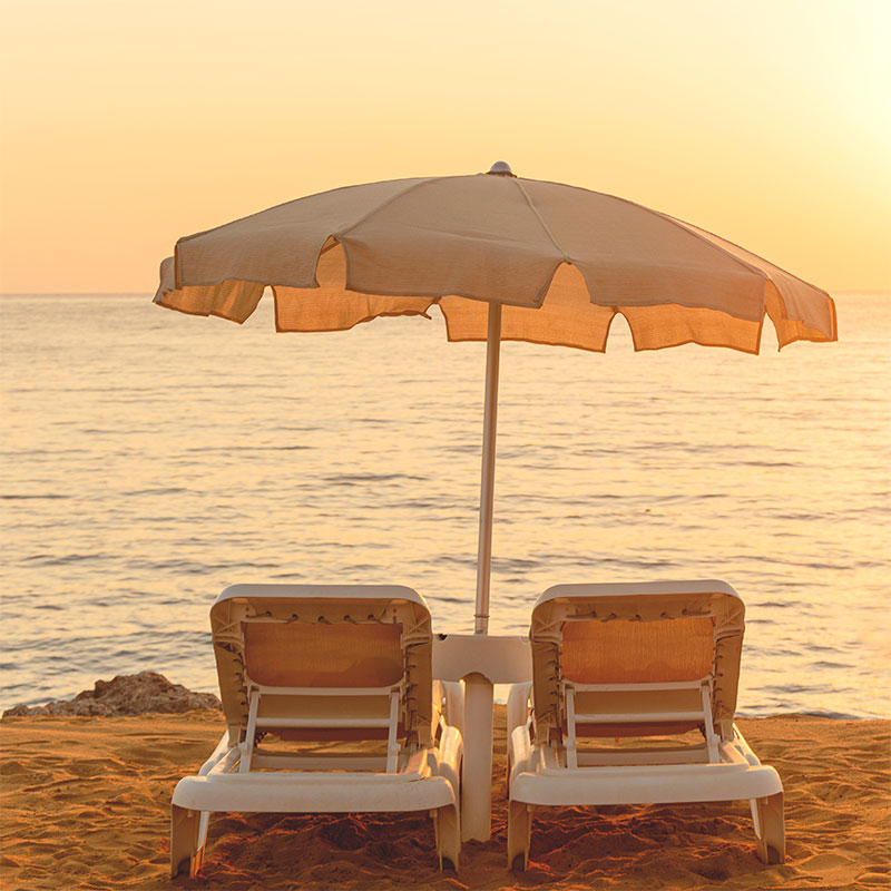 Fotografía de dos hamacas y una sombrilla en una playa con el mar de fondo y el sol al atardecer