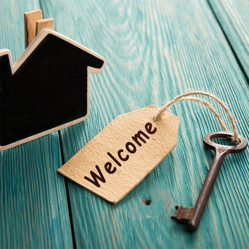 Fotografía de una llave con cartel con la palabra "Welcome" y una pequeña casa de pizarra sobre fondo de madera azul