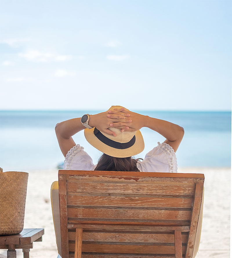 Fotografía de una mujer con un gorro tumbada en una hamaca de madera en la playa observando el mar
