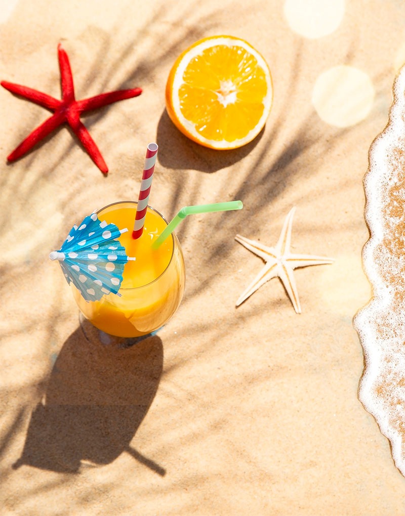 Fotografía de un zumo en una copa con pajitas, media naranja, y dos estrellas de mar en una orilla de playa