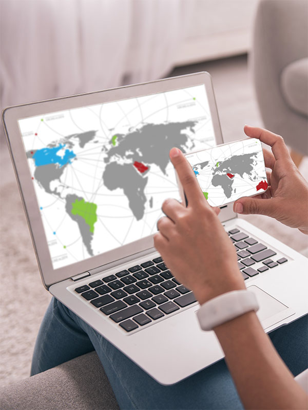 Fotografía de manos de mujer sujetando un móvil que enfoca a una pantalla de ordenador portátil con un mapa del mundo